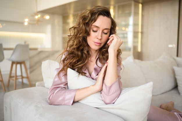 Hermosa mujer joven con pelo rizado en pijama rosa sentado en el sofá blanco o sofá en la mañana. Salón de estilo escandinavo y cocina interior.