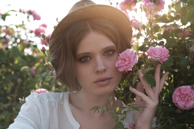 Hermosa mujer joven con pelo rizado posando cerca de rosas en un jardín. El concepto de publicidad de perfumes.