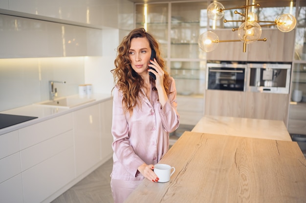 Hermosa mujer joven con pelo rizado tomando café en casa en la mañana de fin de semana perezoso en acogedor pijama rosa.