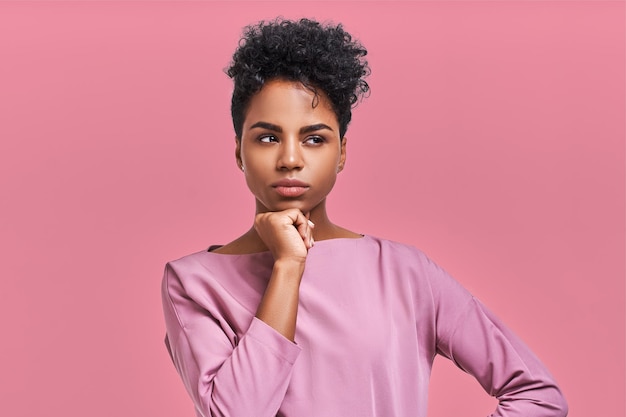 Hermosa mujer joven de piel oscura con peinado afro y aspecto seguro, posa para una revista de moda, mira a un lado cuidadosamente posa contra el fondo rosa del estudio