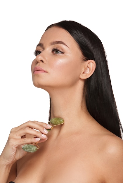 Hermosa mujer joven con una piel perfecta con una toalla en la cabeza con un rodillo facial de jade con piedras de cuarzo natural en blanco