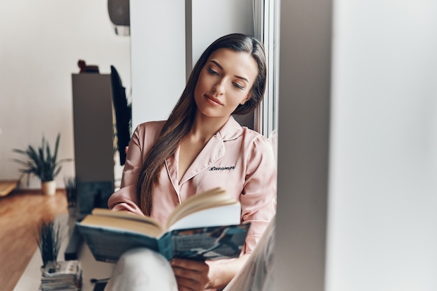 Hermosa mujer joven en pijama acogedor leyendo un libro mientras descansa en el alféizar de la ventana en casa