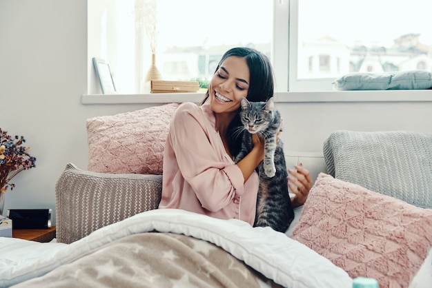 Hermosa mujer joven en pijama sonriendo y abrazando gato doméstico mientras descansa en la cama en casa