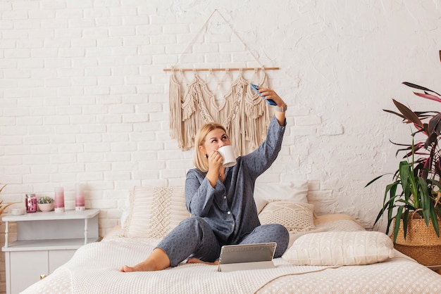 Hermosa mujer joven en pijama trabajando en una computadora portátil en la cama hablando por teléfono Compras en línea