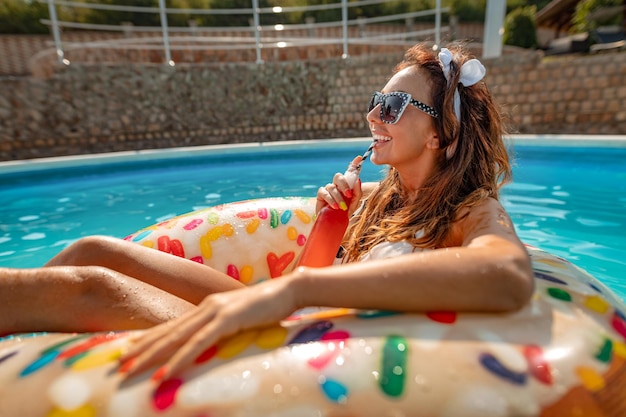 Hermosa mujer joven en la piscina nada en un anillo inflable y se divierte con una botella de cóctel en vacaciones.