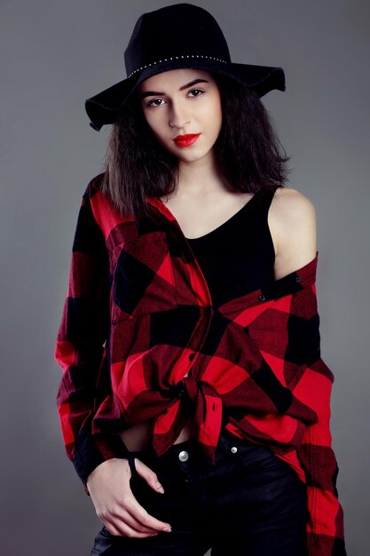 Hermosa mujer joven posando en camisa roja y sombrero negro aislado sobre fondo gris