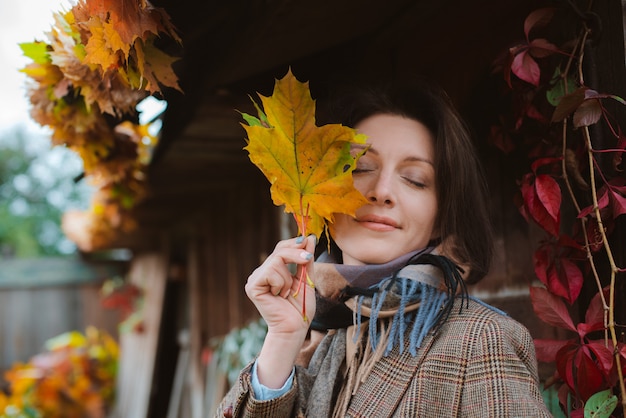 Hermosa mujer joven que cubre su rostro con una hoja amarilla de otoño, sonriendo contra el follaje enrojecido.