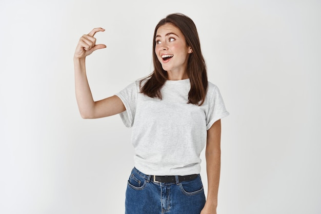 Hermosa mujer joven que muestra un gesto pequeño, minúsculo o pequeño, mirando a la mano con cara sorprendida y feliz, de pie en una camiseta casual sobre una pared blanca.