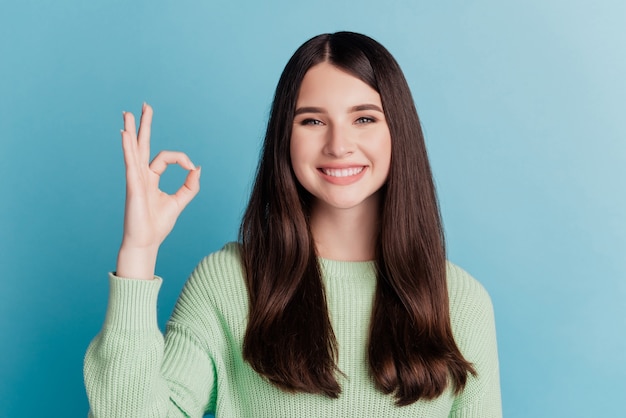 Hermosa mujer joven que muestra la señal de Ok con una sonrisa brillante con dientes en la pared verde azulado