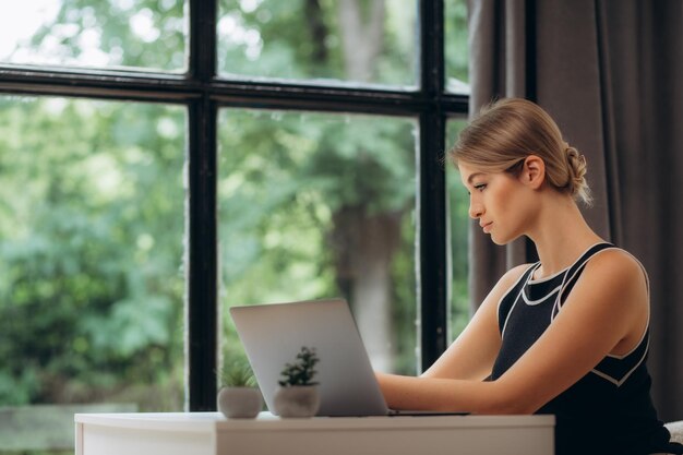 Hermosa mujer joven que trabaja con una computadora portátil en un sofá acogedor cerca de una ventana grande