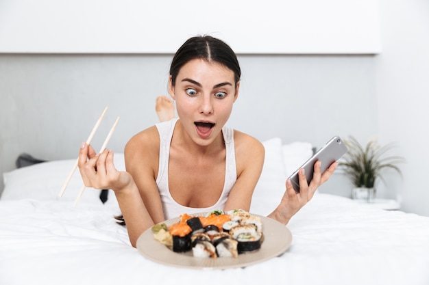 Hermosa mujer joven relajándose en la cama en casa, comiendo sushi de un plato, sosteniendo el teléfono móvil