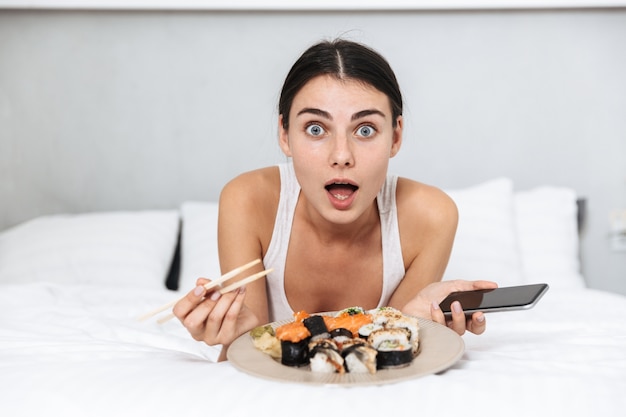 Hermosa mujer joven relajándose en la cama en casa, comiendo sushi de un plato, sosteniendo el teléfono móvil