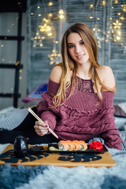 Hermosa mujer joven relajándose en la cama en casa, comiendo sushi de un plato