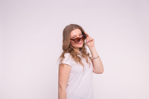 Hermosa mujer joven rizada blanca con camiseta blanca y gafas de sol en la pared blanca con copia