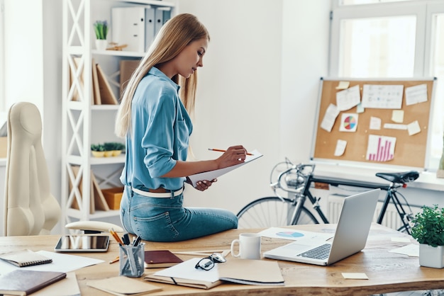 Hermosa mujer joven en ropa casual elegante escribiendo algo y usando la computadora portátil mientras está sentado en la oficina