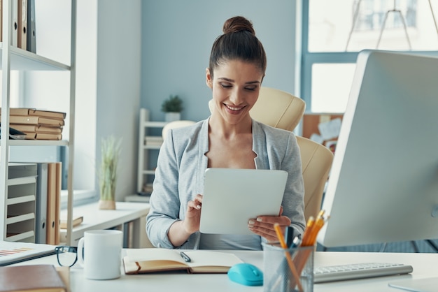 Hermosa mujer joven en ropa casual elegante con tableta digital y sonriendo mientras está sentado en la oficina