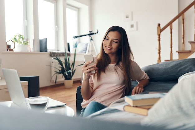 Hermosa mujer joven en ropa casual con teléfono inteligente y sonriendo mientras pasa tiempo en casa