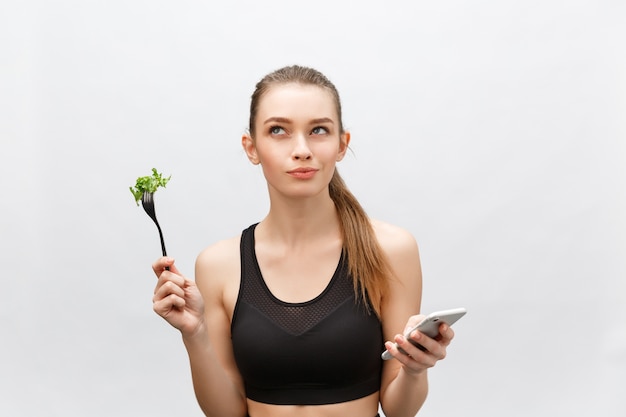 Hermosa mujer joven con ropa deportiva con ensalada en la mano, usando teléfonos inteligentes.