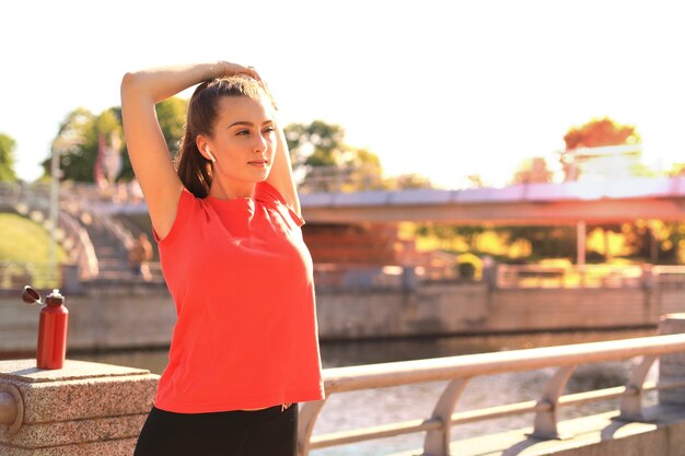 Hermosa mujer joven en ropa deportiva estirando los brazos y mirando concentrada mientras está de pie en el puente.