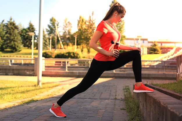 Hermosa mujer joven en ropa deportiva haciendo ejercicios de estiramiento cerca de la orilla del río de la ciudad, sosteniendo una botella de agua.