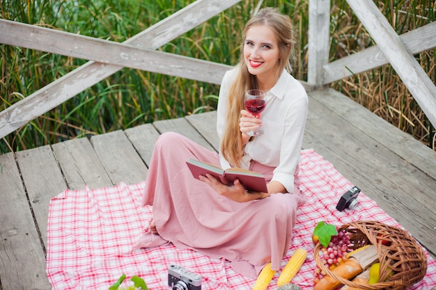 Hermosa mujer joven en ropa vintage tiene picnic en el muelle de madera solo. Picnic estilo francés al aire libre