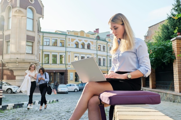 Hermosa mujer joven sentada al aire libre usando el fondo de la calle de la ciudad de la computadora portátil