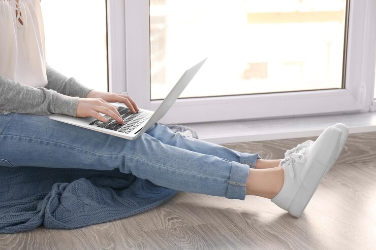 Foto hermosa mujer joven sentada en el alféizar de la ventana y usando una computadora portátil