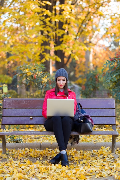 Foto hermosa mujer joven sentada en un banco y usando su computadora portátil en otoño.