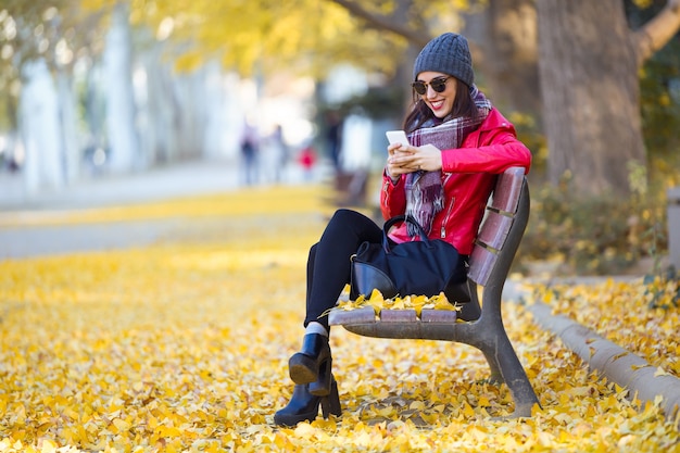 Hermosa mujer joven sentada en un banco y usando su teléfono móvil en otoño.