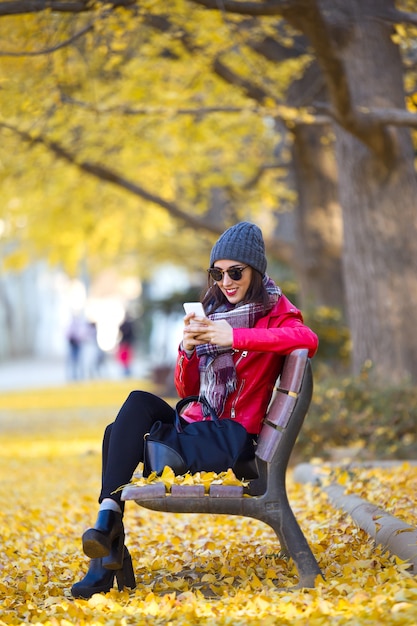 Hermosa mujer joven sentada en un banco y usando su teléfono móvil en otoño.