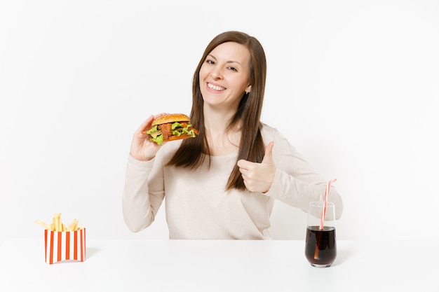 Hermosa mujer joven sentada a la mesa con hamburguesa, papas fritas, cola en botella de vidrio aislada sobre fondo blanco. Nutrición adecuada o comida rápida clásica estadounidense. Área de publicidad con espacio de copia.