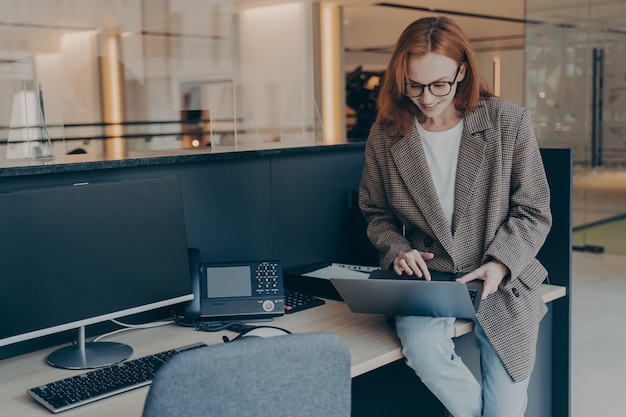 Hermosa mujer joven sentada en la mesa de trabajo en la oficina y usando la computadora portátil