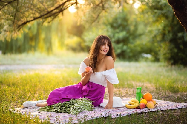 Hermosa mujer joven sentada sobre una manta en la hierba verde y haciendo un picnic