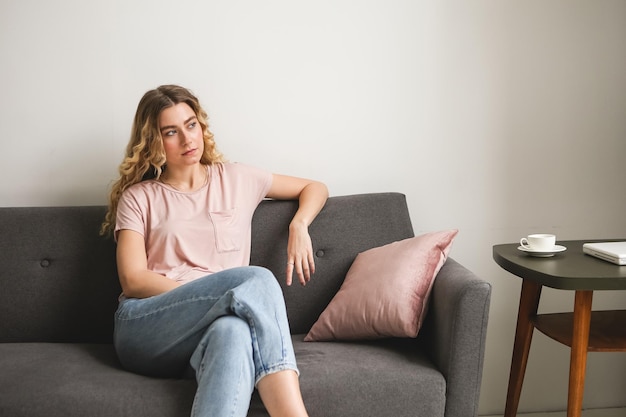 Hermosa mujer joven sentada en un sofá gris en la habitación Chica descansando en camisa rosa Mujer relajante