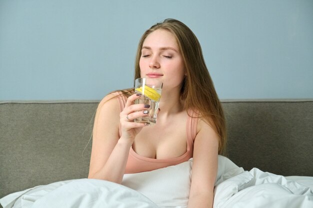 Hermosa mujer joven sentada en su casa en la cama con un vaso de agua con limón por la mañana