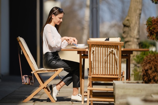 Hermosa mujer joven sentada en la terraza del café en un día soleado y comiendo pastel. Mujer disfrutando del primer sol de primavera al aire libre.