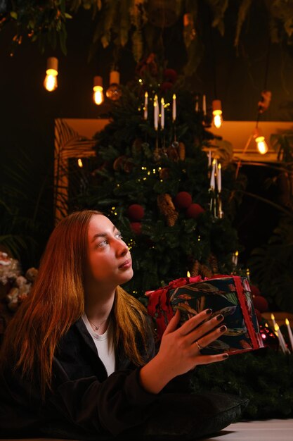 Foto hermosa mujer joven sentada en un traje negro con pestañas blancas al lado del árbol de navidad sosteniendo regalos con franja roja y arco en fondo oscuro