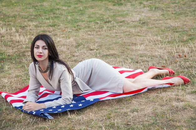Hermosa mujer joven sexy con vestido clásico acostado sobre la bandera americana en el parque. modelo de moda abrazándonos sonriendo y mirando a la cámara. Estilo de vida de Estados Unidos con una gran sonrisa.