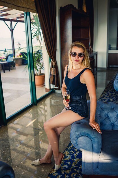 Hermosa mujer joven se sienta en un sillón suave en el vestíbulo del hotel con una copa de champán de vacaciones Lleva gafas de sol y una sonrisa en la cara Concepto de vacaciones