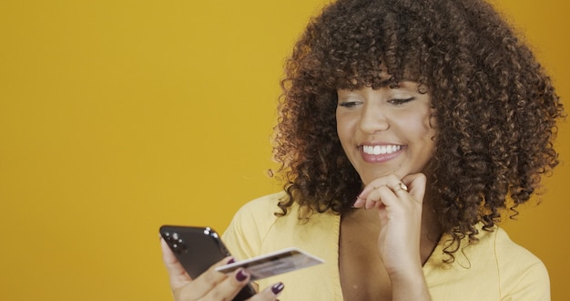 Hermosa mujer joven con smartphone y tarjeta bancaria de crédito. Transacción de compra online. Método de banca móvil sin efectivo. Sonriente joven afroamericana sobre fondo de pared naranja amarillo