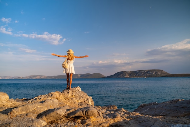 Hermosa mujer joven con sombrero de arrastre de pie en las rocas en vestido blanco junto a la orilla del mar