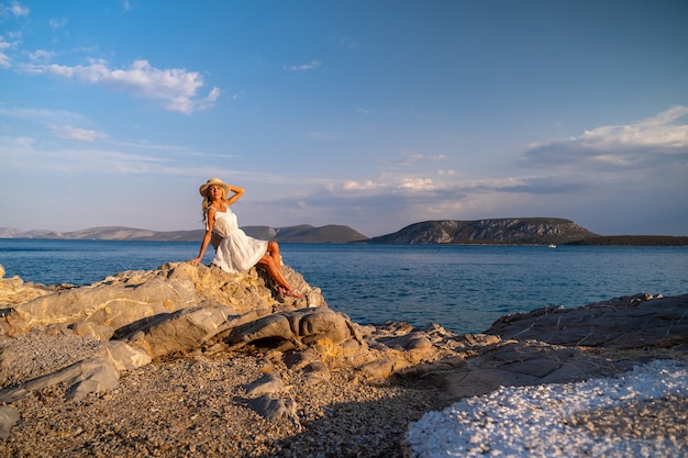 Hermosa mujer joven con sombrero de arrastre sentado en las rocas en vestido blanco junto a la orilla del mar