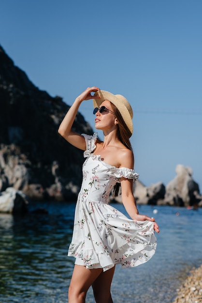 Una hermosa mujer joven con sombrero, gafas y un vestido ligero está caminando por la orilla del océano con el telón de fondo de enormes rocas en un día soleado. Turismo y viajes turísticos.