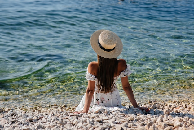 Una hermosa mujer joven con sombrero, gafas y un vestido ligero está sentada de espaldas al océano con el telón de fondo de enormes rocas en un día soleado. Turismo y viajes turísticos.