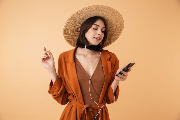 Hermosa mujer joven con sombrero de paja y traje de verano que se encuentran aisladas sobre la pared beige, escuchando música con auriculares y teléfono móvil