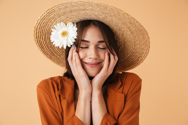 Hermosa mujer joven con sombrero de paja y traje de verano que se encuentran aisladas sobre pared beige, posando con flor de manzanilla