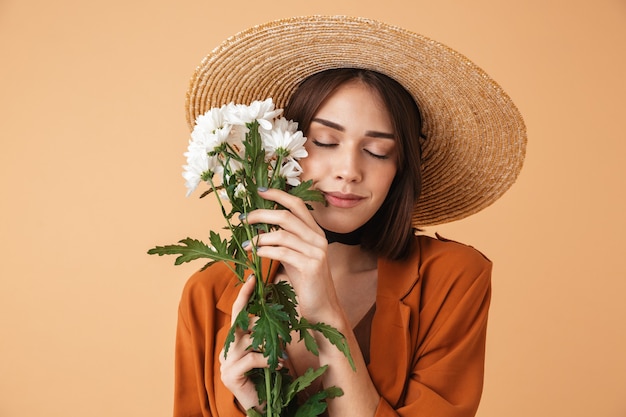 Hermosa mujer joven con sombrero de paja y traje de verano que se encuentran aisladas sobre la pared beige, sosteniendo el ramo de manzanilla