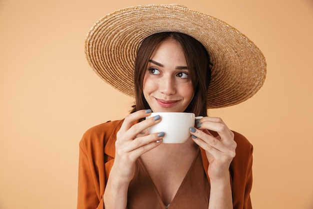 Hermosa mujer joven con sombrero de paja y traje de verano que se encuentran aisladas sobre pared beige, sosteniendo una taza