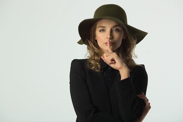 Hermosa mujer joven con un sombrero sobre una pared blanca con un gesto de la mano que significa silencio