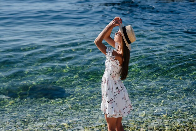 Una hermosa mujer joven con un sombrero y un vestido ligero de espaldas está caminando a lo largo de la orilla del océano con el telón de fondo de enormes rocas en un día soleado. Viajes de turismo y vacaciones.